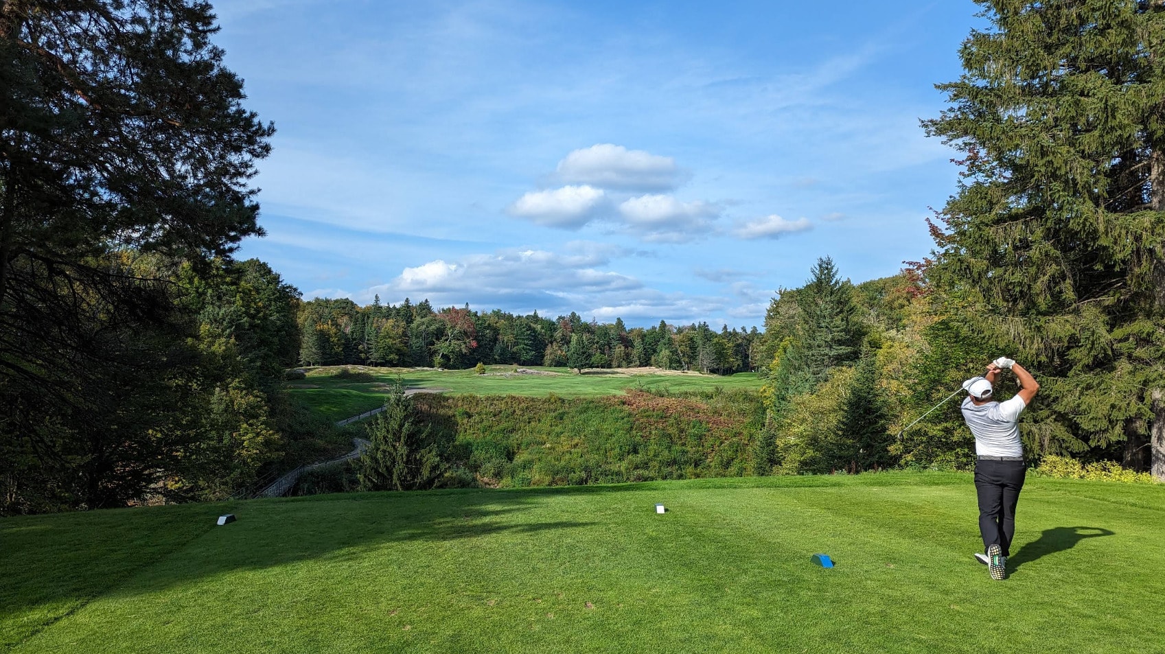A golfer tees off at Club de Golf Grand-Mère, surrounded by lush forest and rolling fairways under a bright blue sky. A golfer tees off at Club de Golf Grand-Mère, surrounded by lush forest and rolling fairways under a bright blue sky.